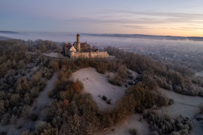 Die Altenburg an einem frostigen Wintermorgen um die Sonnenwende, Raureif auf dem Hangwald, Bamberg rechts im Dunst des aufgehenden Tages. 21. Dezember 2021.