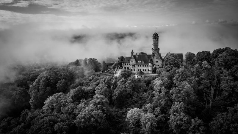 Die Burg über der Nebeldecke unter dramatischem Wolkenhimmel. Schwarzweiße Bearbeitung einer eigenen Drohnenaufnahme.